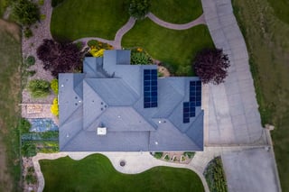 Aerial view of a custom home with gray roof, solar panels, and curved driveway surrounded by landscaped yard.