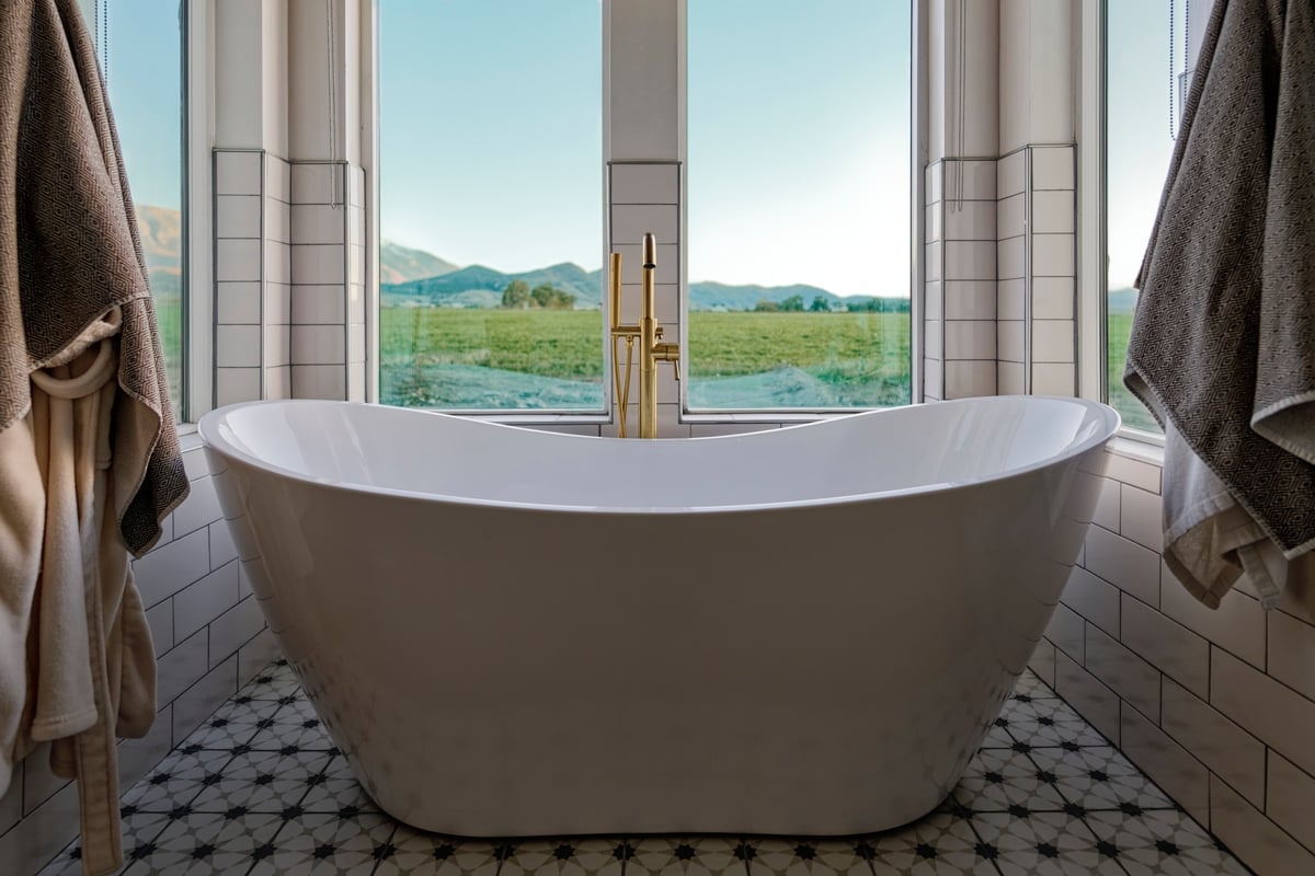Modern custom home bathroom with white freestanding tub, gold faucet, and scenic mountain view.
