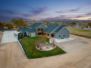 Spacious custom home at dusk with glowing windows, curved lawn, and stone fire pit in rural setting.