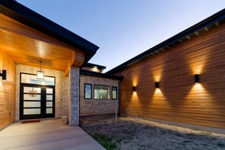 Modern custom home entrance at twilight with warm lighting, stone accents, and wood siding.