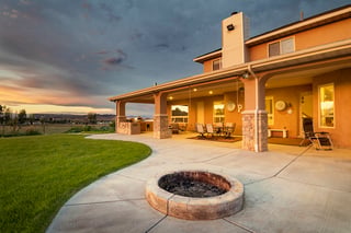 Custom home backyard with covered patio, stone fire pit, and sunset sky over open landscape.