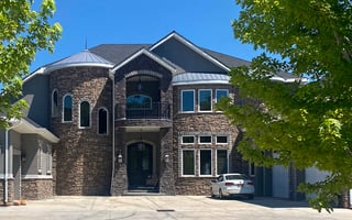 Luxury custom home with stone exterior, turret, balcony, and parked sedan under a clear blue sky.