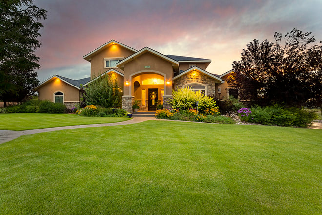 Elegant custom home at dusk with glowing entrance, manicured lawn, and colorful garden under twilight sky.