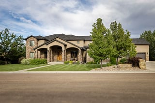 Spacious custom home with stone and stucco exterior, landscaped lawn, trees, and driveway under bright sky.